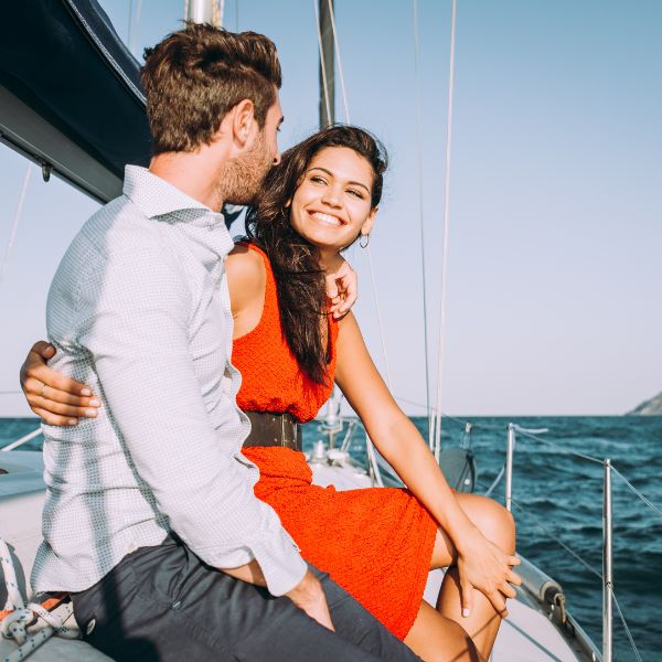 A romantic scene on a boat showcasing a stylishly dressed couple with the man in a grey shirt and the woman in a vibrant red dress.