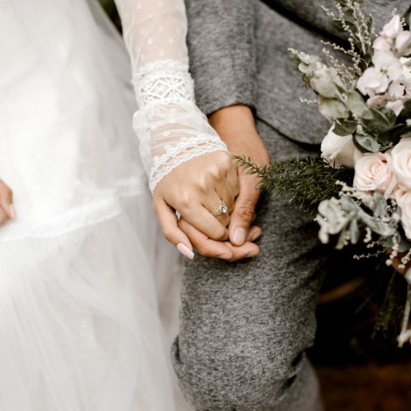 A newlywed couple holding hands, with the bride wearing a white lace dress and a sparkling ring