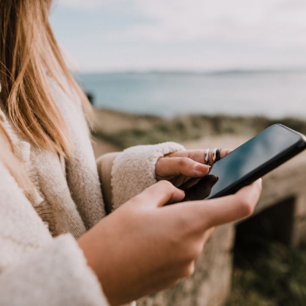 A person in a cozy sweater using a smartphone with a scenic coastline in the background, captured from over the shoulder.