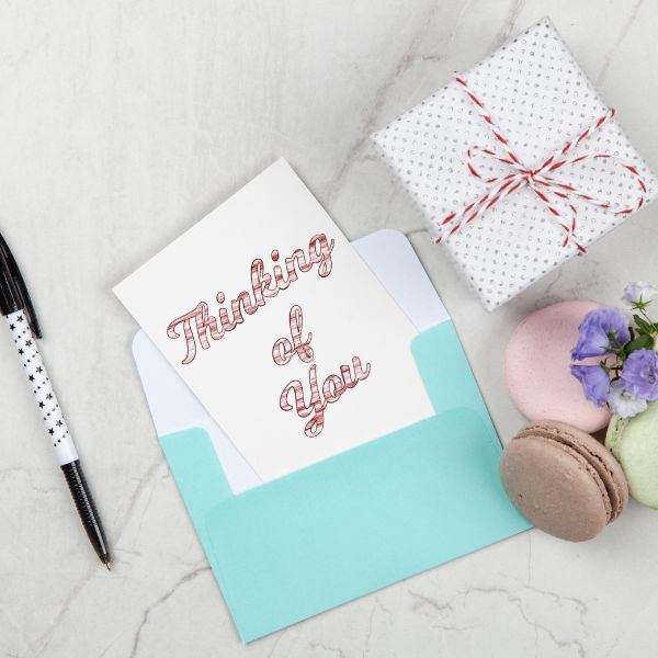 A neatly arranged greeting card with "Thinking of You" written in cursive, accompanied by a small gift box, macarons, and flowers on a marble surface