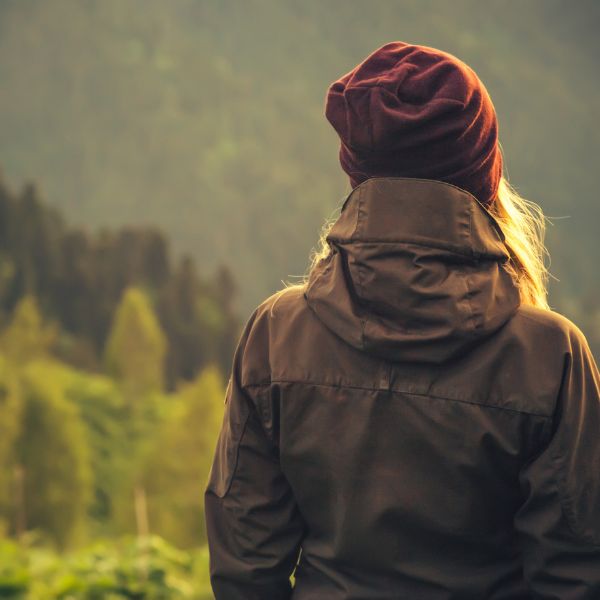 The back view of a person wearing a maroon beanie and brown jacket, gazing into a lush green forest.