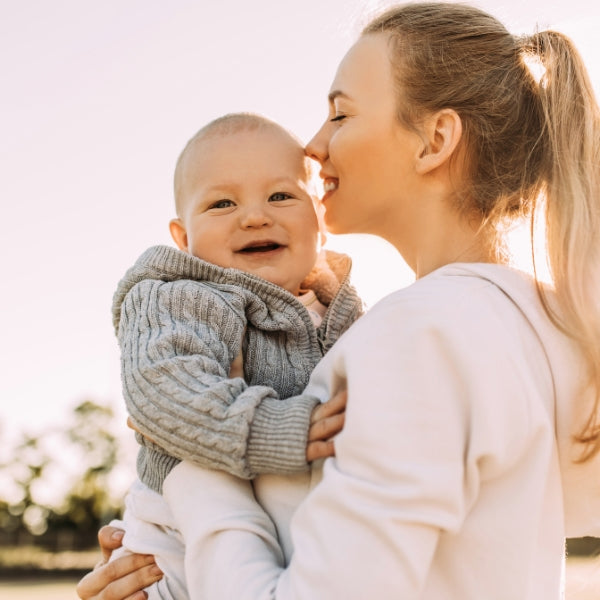 Gleeful baby in a cozy sweater being kissed by a smiling mother outdoors, radiating the joy of a happy first Mother's Day.