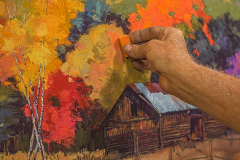 A hand with a piece of chalk pastel adding orange shading to a cabin in fall surrounded by foliage