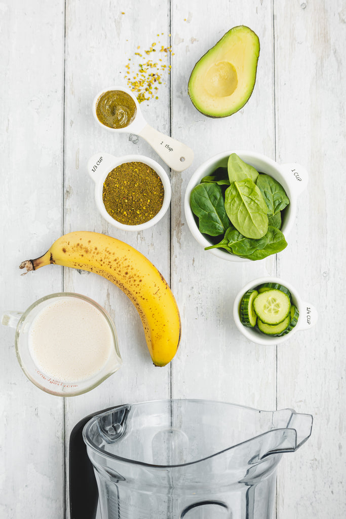 avocado, pistachio pieces, spinach, cucumber, banana, and soy milk on light white wood backdrop from overhead, with blender