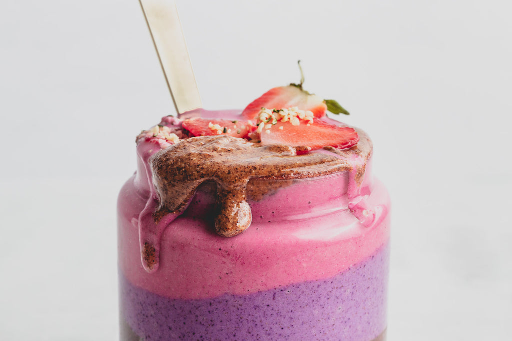 close up of pink and purple smoothie in glass jar with hazelnut butter, strawberries, and hemp seeds on top