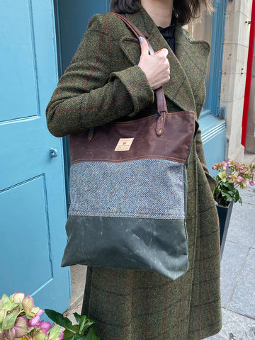 Image features a tote bag worn over the shoulder by a model wearing a green tweed coat. The tote bag is made from a rust waxed cotton, with a Harris Tweed panel in the middle and an olive waxed cotton on the bottom