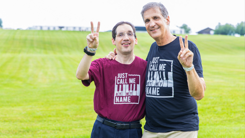 Ken Trush standing next to his son Daniel Trush in an open field at Woodstock. They are wearing T-shirts which say "Just Call Me By My Name"