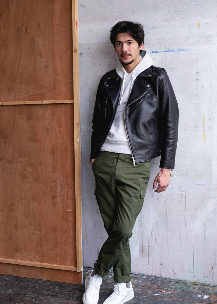 Homme debout en hoodie gris, blouson perfecto noir et pantalon cargo vert.