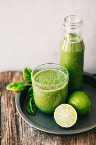 a-glass-and-a-bottle-with-green-juice-on-a-plate-with-green-lime-over-wodden-table