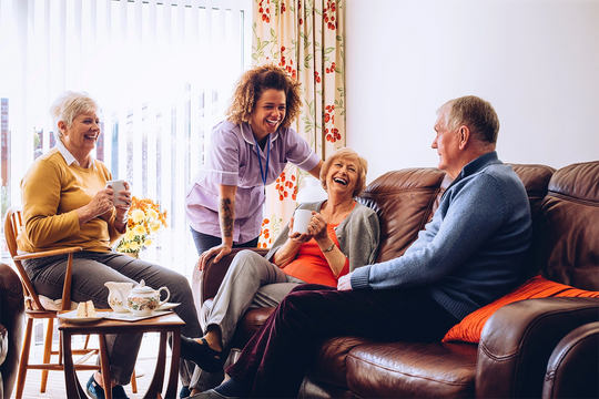 a female carer speaking to two female and one male patient with a cup of tea and cake in a care home setting