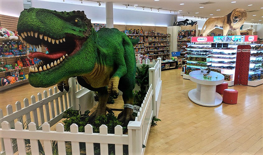 14 foot green T-Rex in a fenced in area and a life-sized lion that sits on top of a gondola of Schleich products, which is located at Maziply Toys, the US flagship store for Schleich.