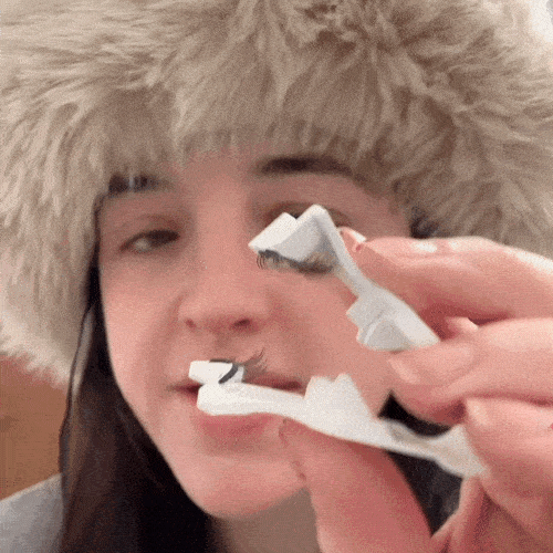 Person wearing a fur hat, showcasing and applying false lashes with a plastic applicator.