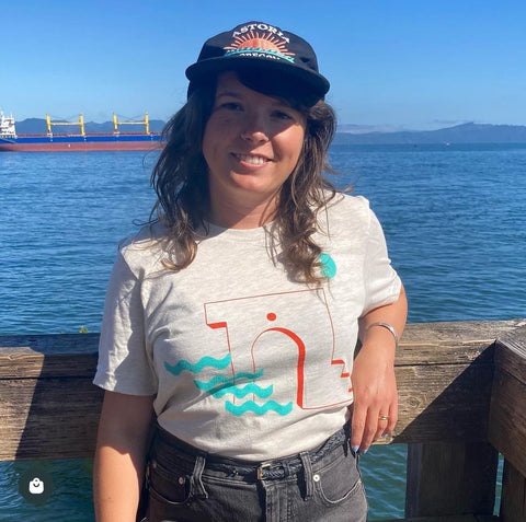 A photo of a woman in her early 30s with long brown hair wearing a black hat that says "Astoria, Oregon" and a shirt with a turquoise and terracotta abstract stairway design. She is smiling and standing in front of the river with a cargo ship in the background.