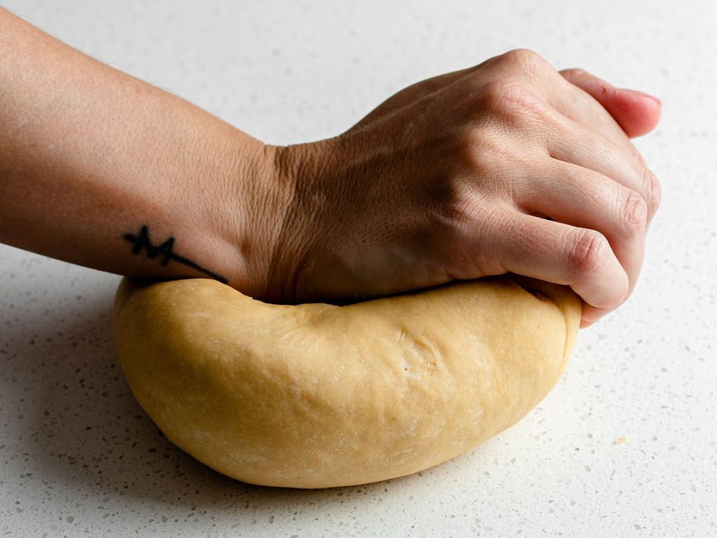 A hand is kneading pasta dough on a white speckled surface. The dough is pale yellow and smooth.