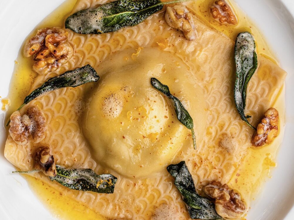 Bird's eye view of a giant square raviolo resting on a white plate. We see an archway pattern on the edges of the ravioli, and a plump center. The raviolo is covered in a brown butter sauce (golden in color) and sprinkled with whole fried sage leaves and walnuts. 