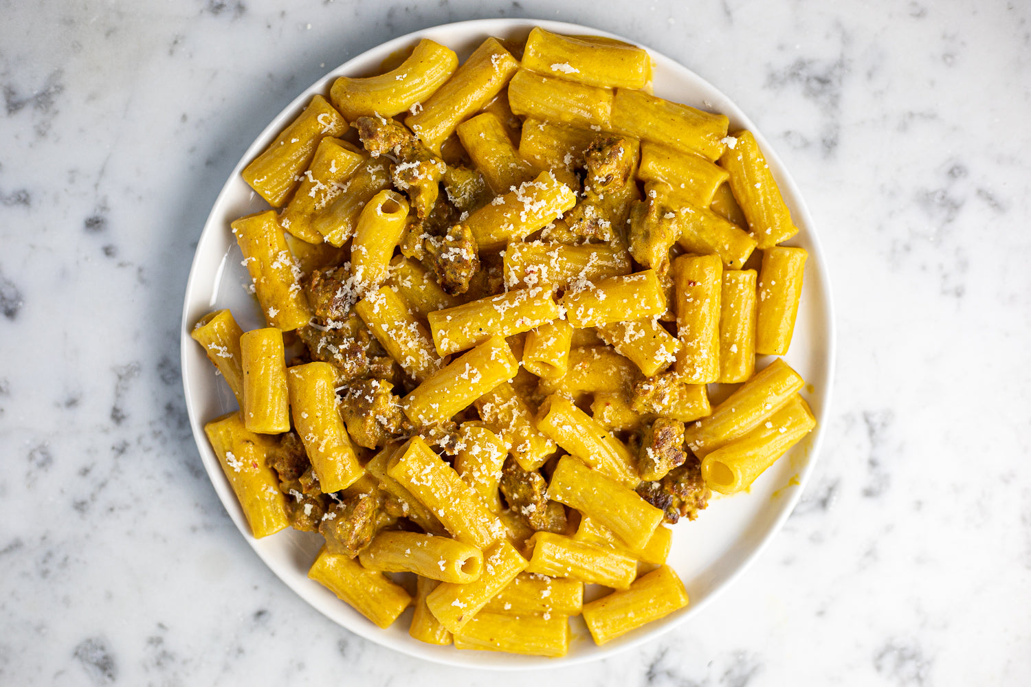 A plate of yellow colored tube pasta with crumbled bits of brown sausage. A light sprinkling of white parmesan cheese is on top.