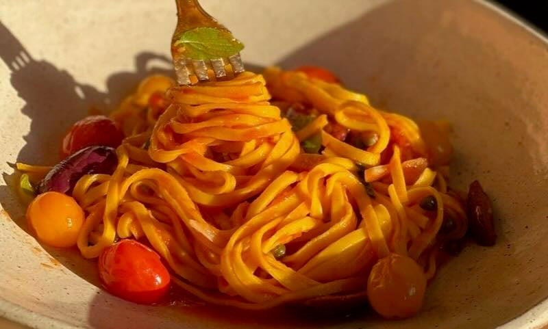 We see a bowl of spaghetti coated in a red tomato sauce. There are a few orange and red cherry tomatoes resting around the spaghetti. A warm sunlight hits the pasta dish, creating dramatic shadows. There is a fork dipping into the spaghetti.