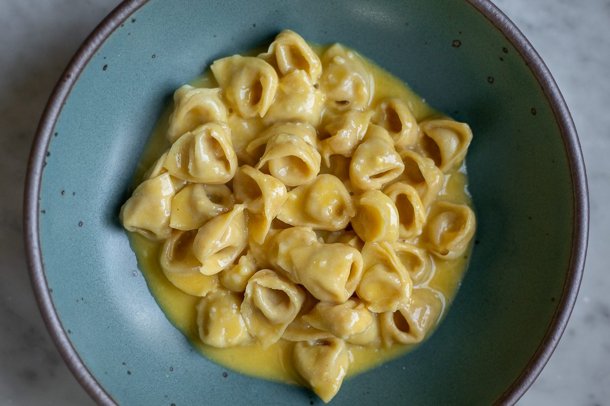 A teal low-bowl holds a heaping of golden tortellini pasta with a creamy yellow sauce. 