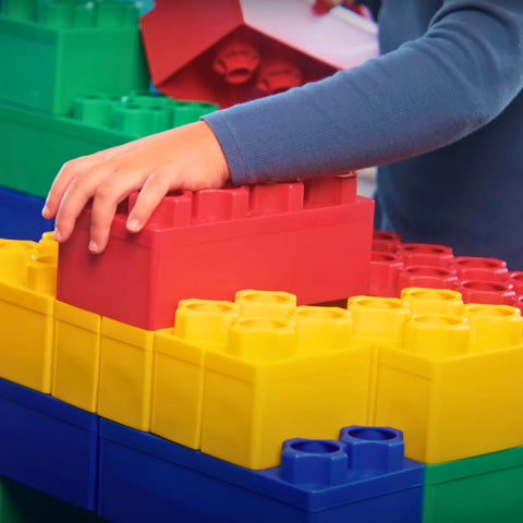 A child's hand places a large red Biggo Block onto a colorful structure made of yellow, blue, and green jumbo blocks, highlighting the ease and fun of building with oversized pieces.