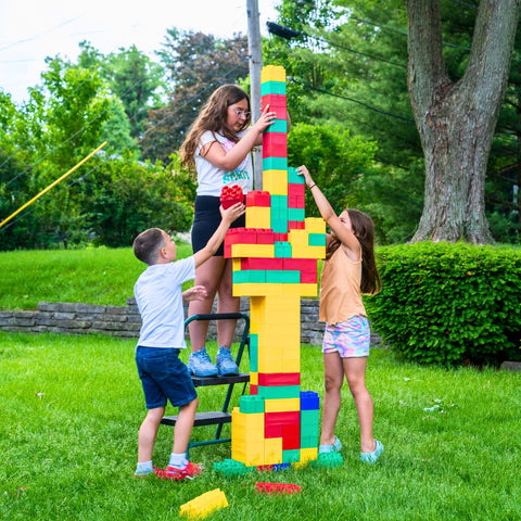 Children work together to build a tall, colorful structure with extra-large building blocks, showcasing teamwork and creativity in an outdoor setting.