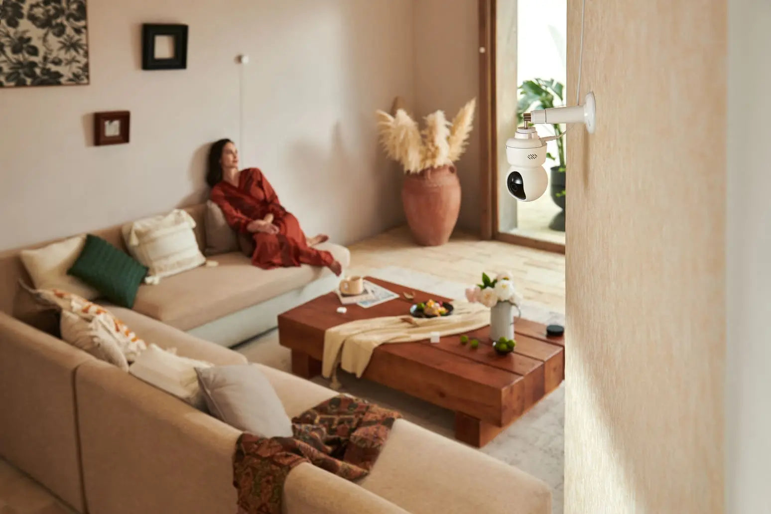 A girl in red loungewear is reclining on the living room sofa, with a camera E1 mounted on the wall not far away.