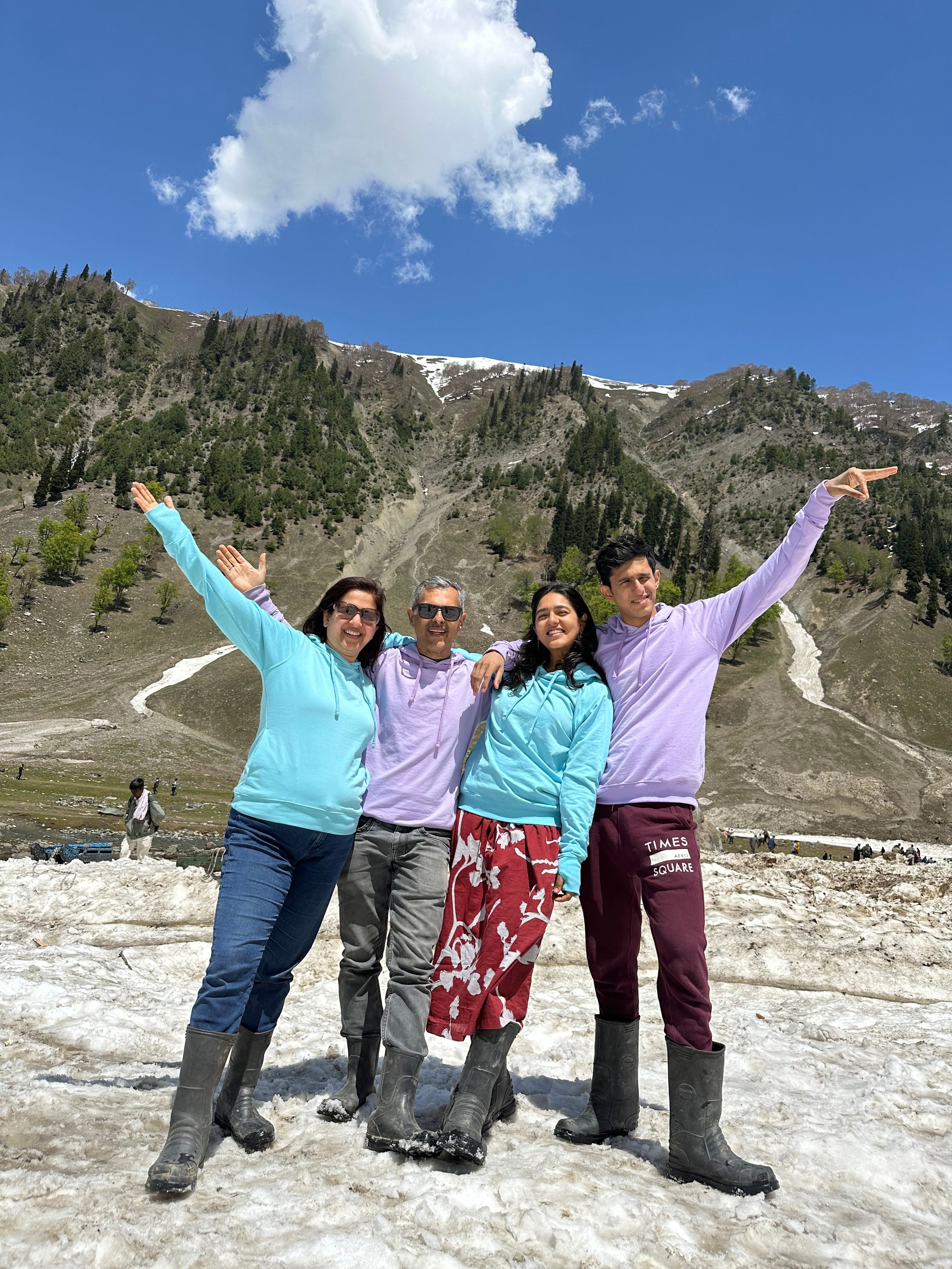 A smiling family of four standing on a snowy slope with lush green mountains in the background, all dressed in pastel-colored hoodies and rubber boots, arms raised in celebration.