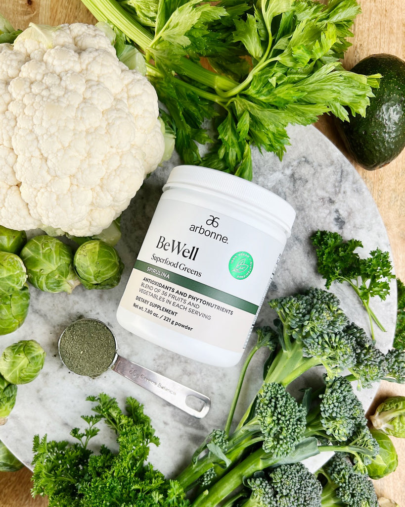 Top view of Arbonne BeWell Superfood Greens on a marble surface, accompanied by an assortment of green vegetables like cauliflower, celery, avocado, and broccoli.