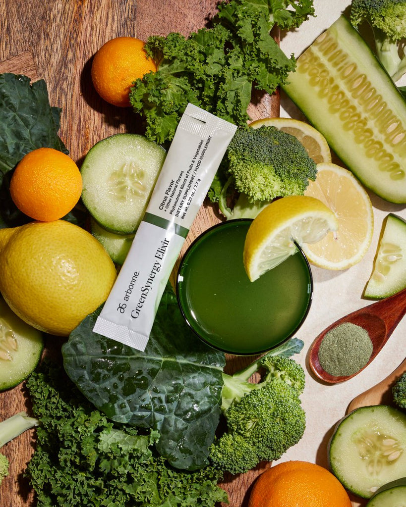 Arbonne GreenSynergy Elixir packet surrounded by fresh citrus fruits, cucumbers, kale, broccoli, and a green drink on a wooden surface.