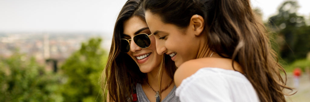 Deux femmes joyeuses portant des lunettes de soleil partageant un moment proche, souriant et s'embrassant à l'extérieur avec un fond de paysage urbain flou, transmettant bonheur et amitié