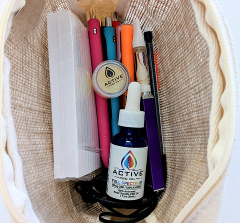 The contents of a hemp bag are shown with pink, blue, and red pens, an orange highlighter, a black mechanical pencil, a pack of index cards wrapped in plastic, a purple vape battery with an oil filled cartridge attached, a blue 1 oz glass bottle with a white label with rainbow lettering stating "Active CBD Oil Full Spectrum," a quarter sized jar of a cream colored salve, and wired headphones tucked in the bottom of the bag.