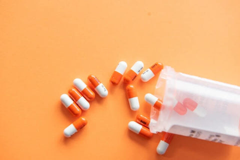 A clear pill bottle with a white label is shown laying down on a light orange background with twelve orange and white pills spilled on the background and three orange and white pills remaining in the bottle.