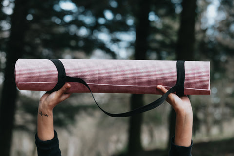 A person's hands are shown holding a pink yoga mat with black handles above their head while in front of some trees.