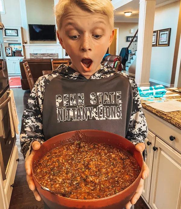 Surprised young boy holding a large bowl of salsa in a kitchen.