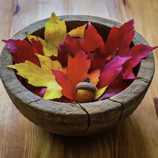 Rustic wooden bowl overflowing with colorful fall leaves, pinecones, and acorns on a cozy knit blanket.