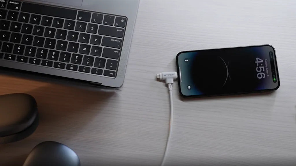 Eine Laptop-Tastatur und ein mit einem Ladekabel verbundenes Smartphone auf einer hellen Holzoberfläche.