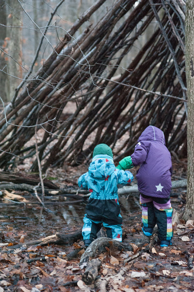 two kids in the woods at a wood teepee in waterproof winter gear from biddleandbop