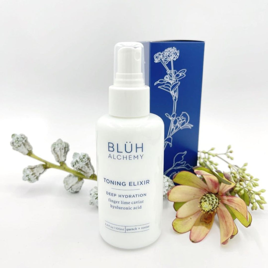 On the photo is a white bottle of a skincare product. In blue color it writes Bluh Alchemy Toning Elixir deep hydration finger lime caviar hyaluronic acid. This is a face mist. Next to the product is a flower and some more small plants. Behind is product's paper box - blue with white drawing of a plant.