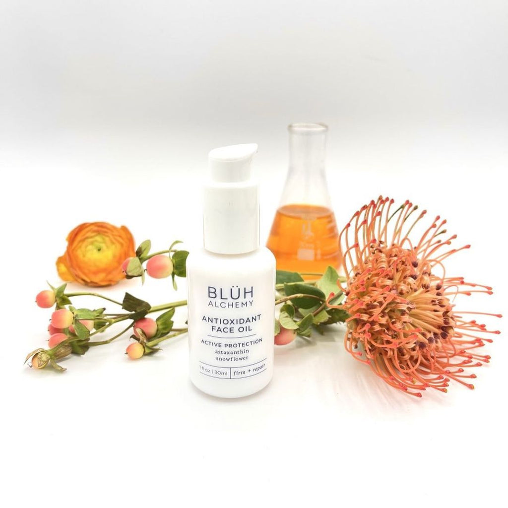 On the photo is a skincare product from a brand Bluh Alchemy. It is their Antioxidant Face Oil. White packaging, white glass bottle with a pump. Writings on the bottle are in blue. There are also three different flowers in this photo and a vail with this oil which has a rich deep orange color.