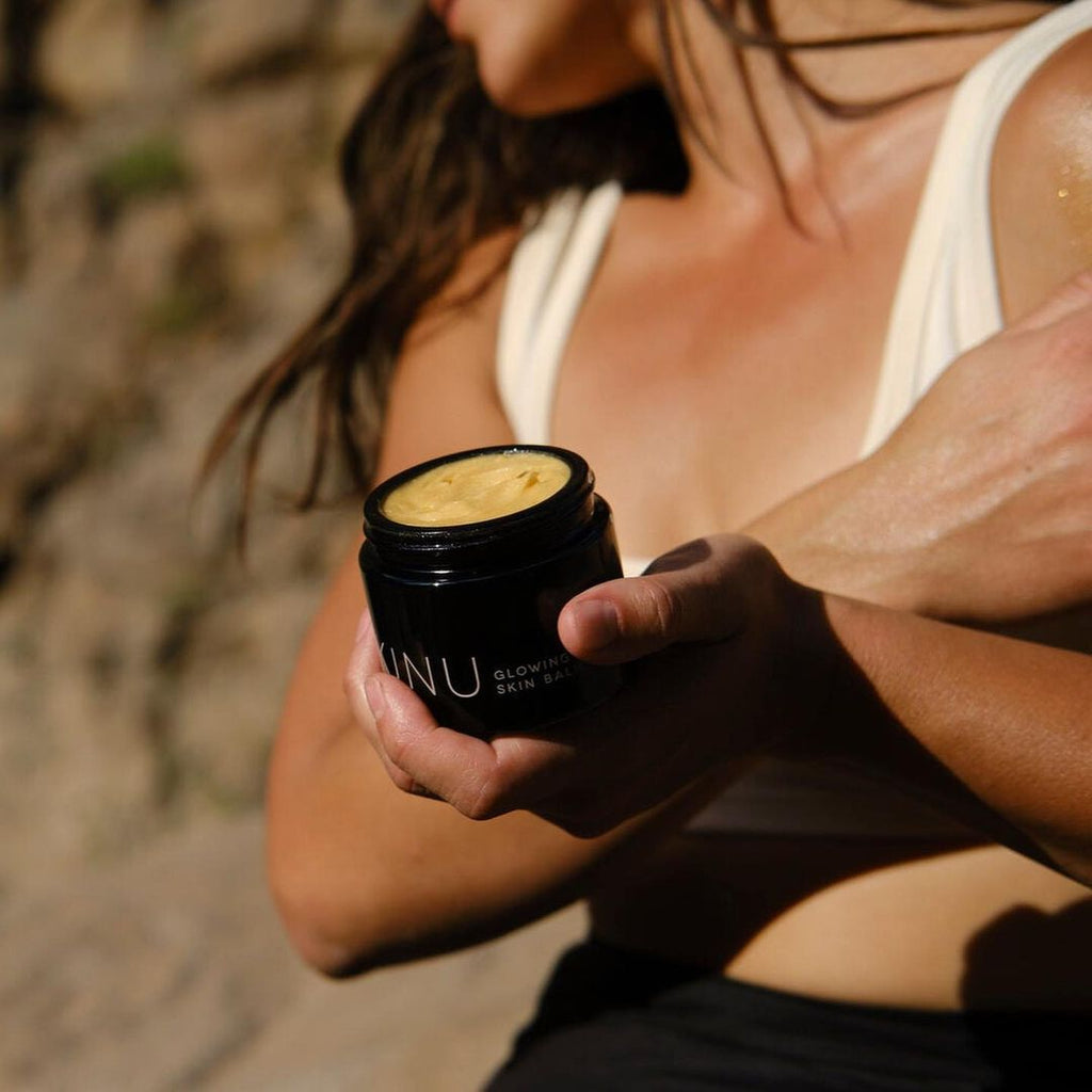 a woman is holding a black jar of a body butter with yellow color. This is the H si for Love KINU glowing skin balm