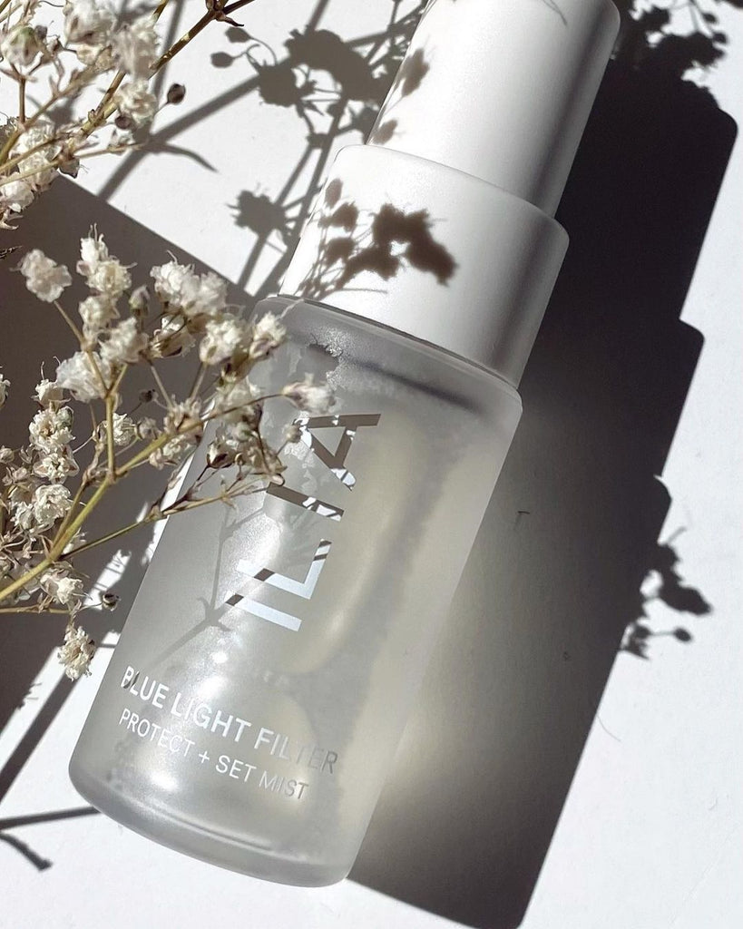 A close up of a face mist in frosted clear glass packaging with white spray mister. It lays on a white background and over it is a plant with tiny, delicate flowers in neutral color. The product is a new products from ILIA, the Blue Light Face Mist.