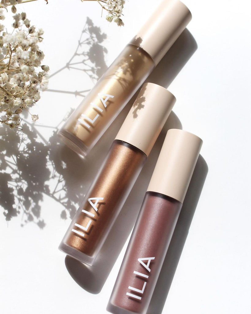 Liquid eyeshadows lay on white background. From top to bottom are: Gleam, an antique gold color, then Sheen, a copper bronze and Dim, Gray, a lavender with pink and purple pearl. These are Eye Tints, liquid eyeshadow that turn from cream to powder, by ILIA Beauty.