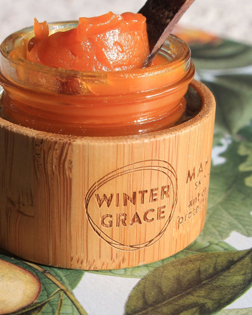 a close up of a bamboo and glass jar skincare product that is open and you can see the vibrant orange color of the product. also a wooden spoon is in the balm to show the texture. This product is the winter grace by mahalo skin care