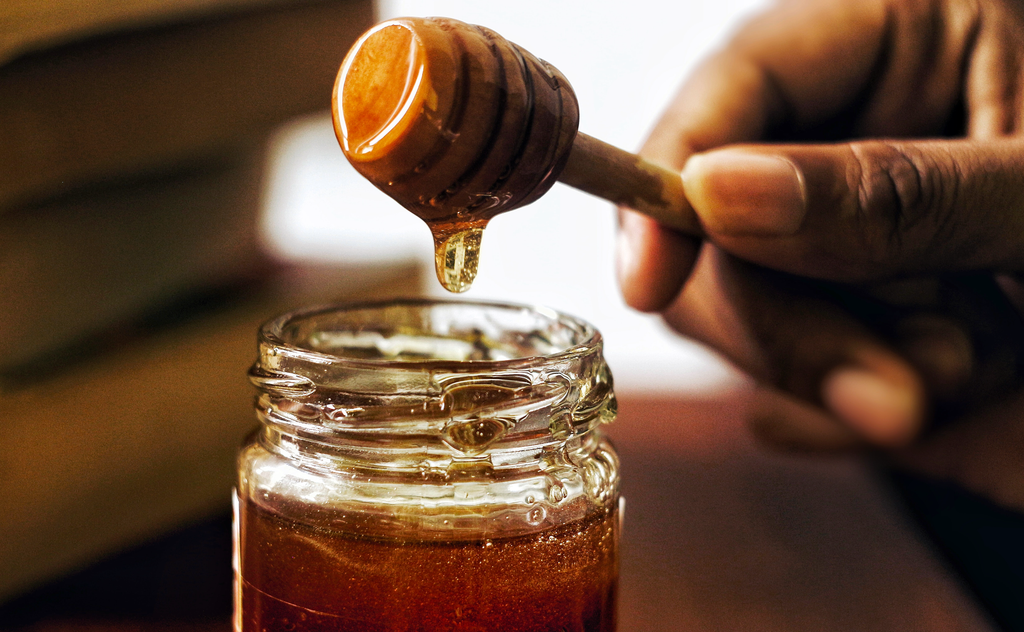 A hand holding a honey dipper over a jar of raw honey