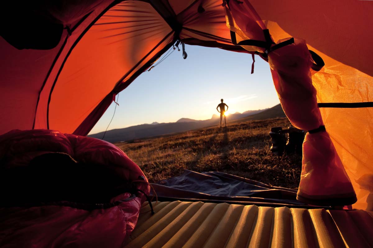 View from inside of a tent with camper standing outside watching the sun set.