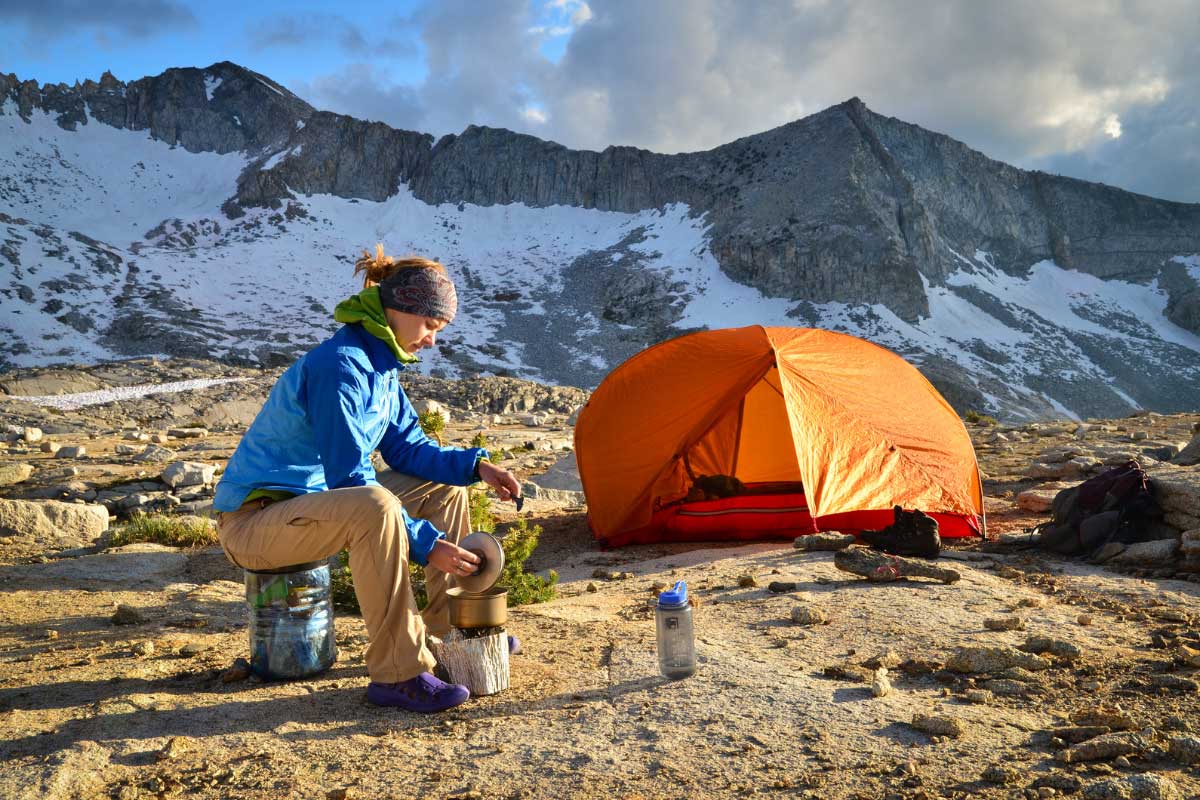 Backpacker sitting near their tent preparing a meal on a small stove with snow covered mountains in the background.