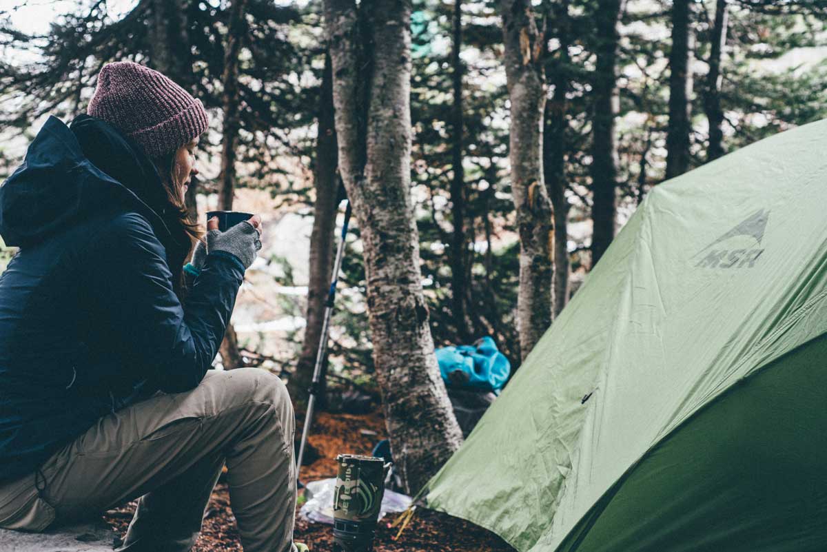 Camper wearing warm layers holding hot coffee next to tent.