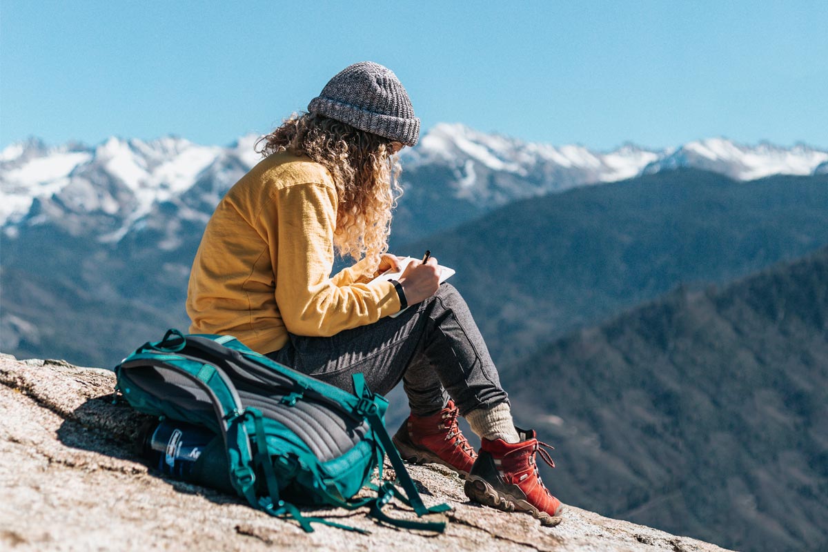 Hiker sitting on a rocky ridge with mountain views in the background and trail journaling