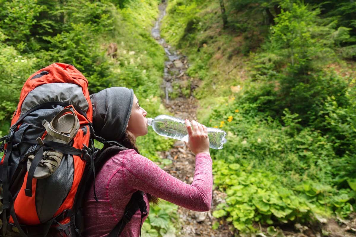 Backpacker taking a drink from water bottle in the woods with a small stream behind.