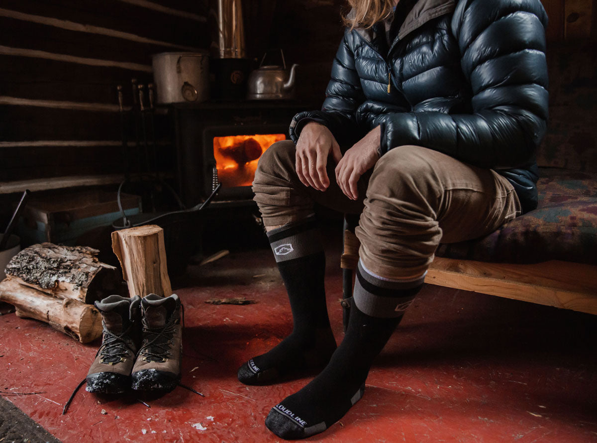 Man warming up near a wood stove wearing Cloudline Apparel merino wool snow socks in a log cabin.