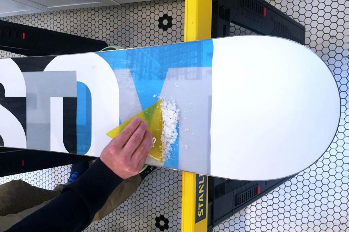 Person using a plastic base scraper to remove excess wax after hot waxing snowboard.